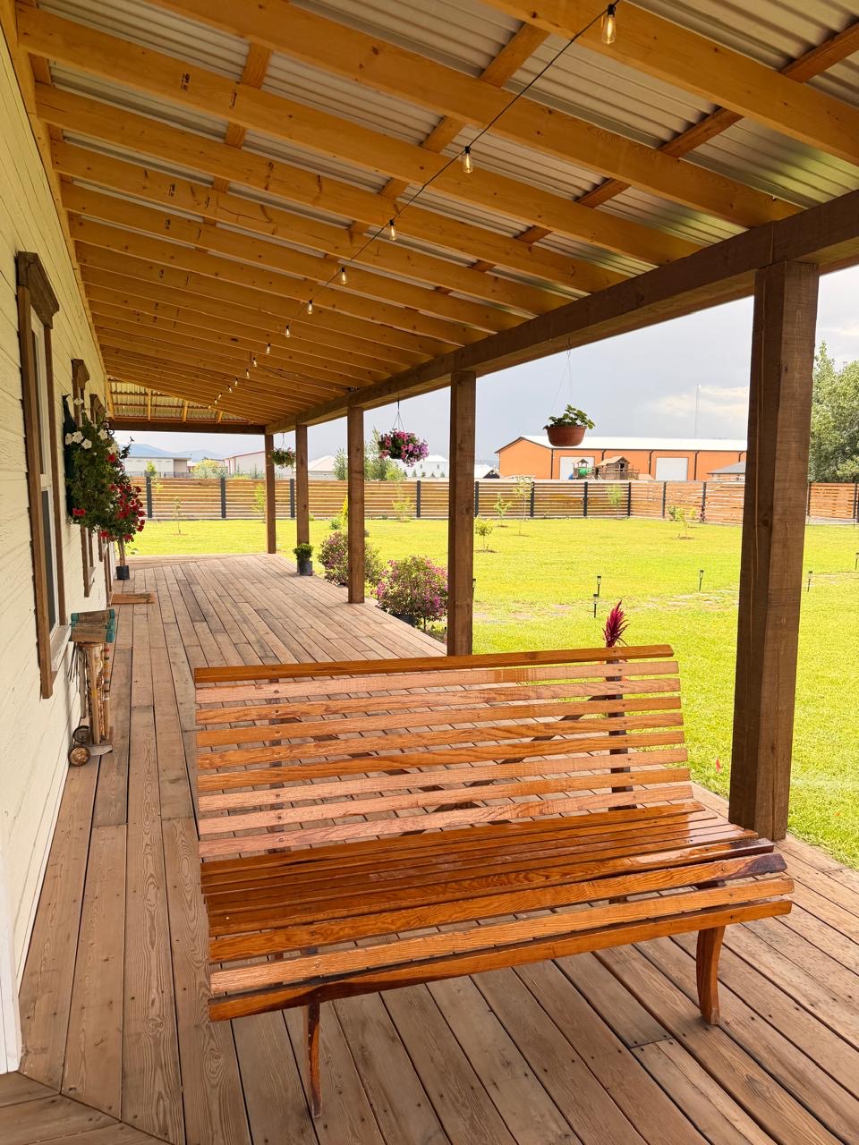 Covered porch with a slatted wooden bench and string lights looking over the yard
